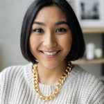 Portrait of a cheerful young woman wearing a gold necklace and sweater indoors with a blurred background.