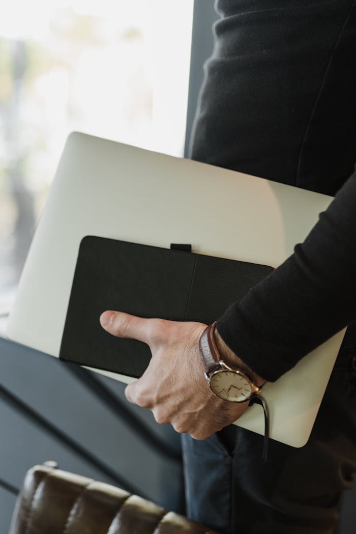 A detailed close-up of a hand holding a laptop and notebook, showcasing a watch.